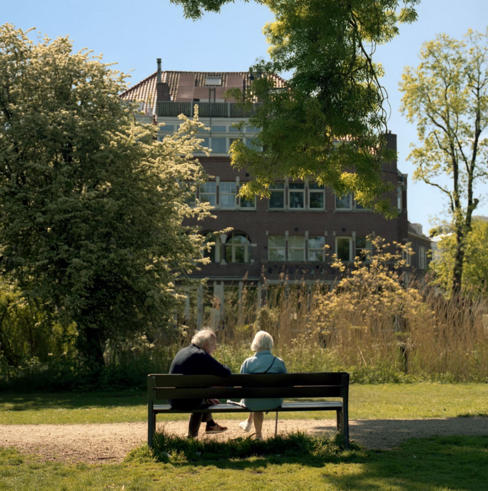 Een ouder echtpaar zit samen op een bankje in een zonnig Nederlands park